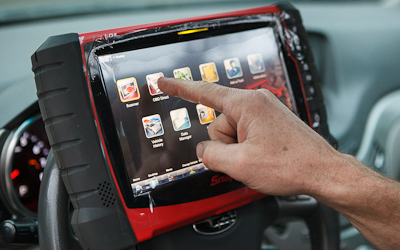 A hand tapping the screen of a handheld diagnostic scanner mounted on a steering wheel